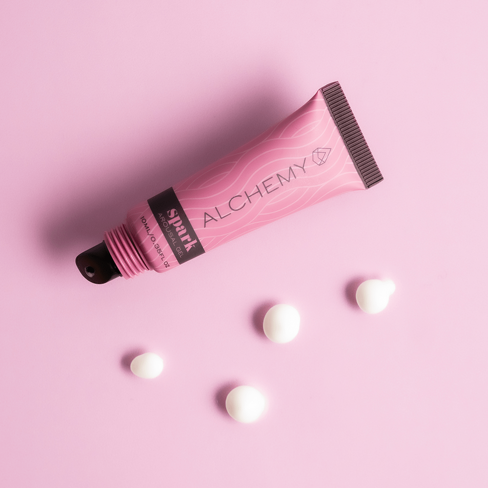 Top view of Alchemy Spark Female Arousal Gel 1oz tube on a pink background with dots of gel surrounding it