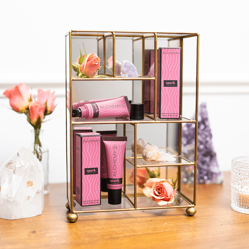Alchemy Spark Female Arousal Gel 1oz tubes and boxes in a compact gold-colored shelf organizer on a wood surface