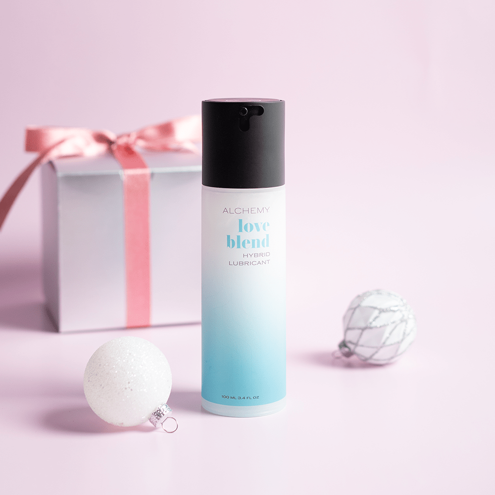 Alchemy Love Blend: Hybrid Personal Lubricant bottle with holiday gift boxes, ornaments, and light pastel background