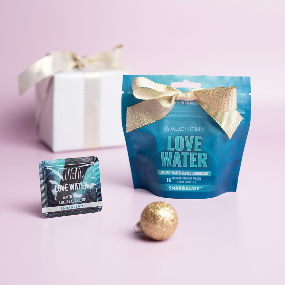 Alchemy Love Water Water-Based Snap + Slide Single-Use Personal Lubricant Packets with holiday gift boxes, ornaments, and light pastel background