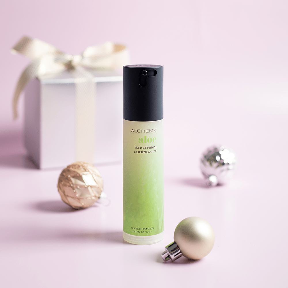 Alchemy Aloe: Hydrating Water-Based Personal Lubricant bottle with holiday gift boxes, ornaments, and light pastel background