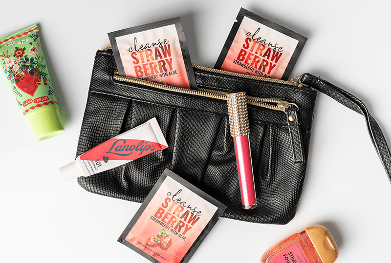Alchemy Cleanse Strawberry Scented Wipes top view with purse and makeup accessories on a white surface