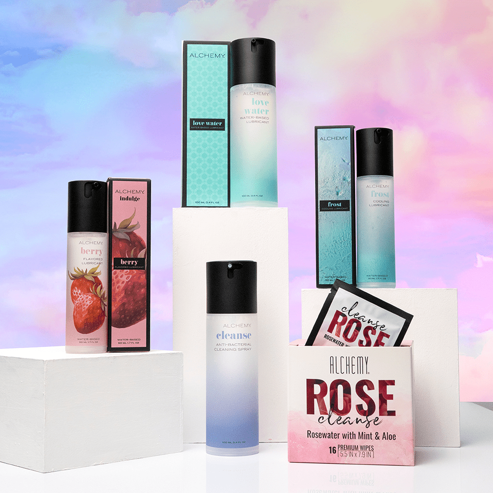 Alchemy Essentials Bundle: Water-based personal lubricant bottles and boxes on white blocks and a colorful cloudy sky background