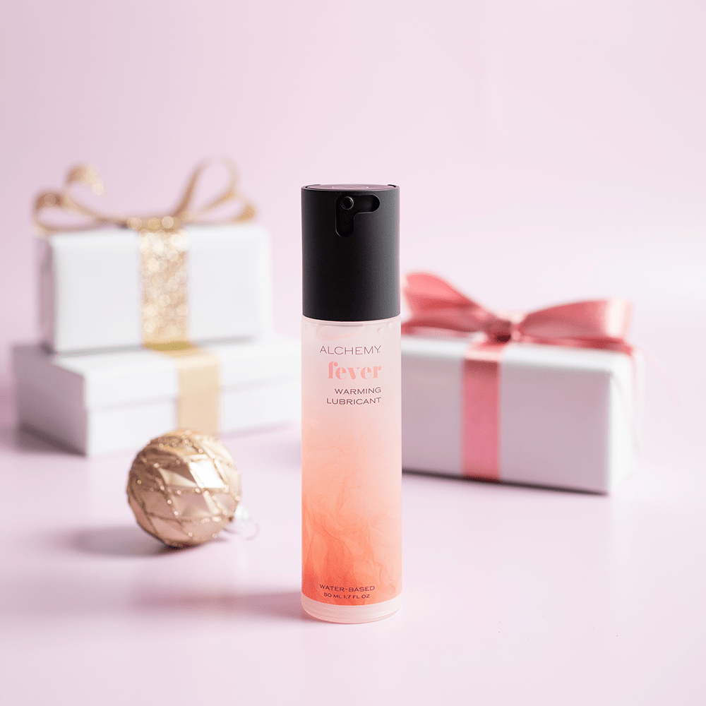 Alchemy Fever: Water-Based Warming Personal Lubricant bottle with holiday gift boxes, ornaments, and light pastel background