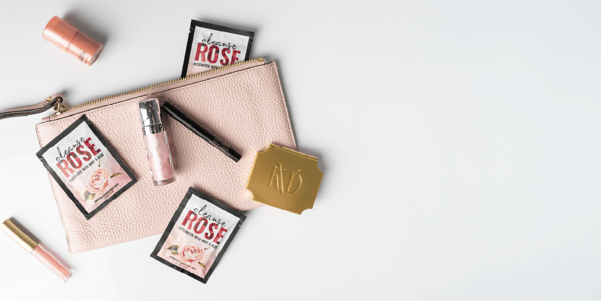 Alchemy Aftercare collection: arrangement of beauty products and Alchemy Cleanse Wipe packets on a white background, featuring a light pink textured zip pouch, two pink lipsticks, a tube of lip gloss, and a black eyeliner pencil.