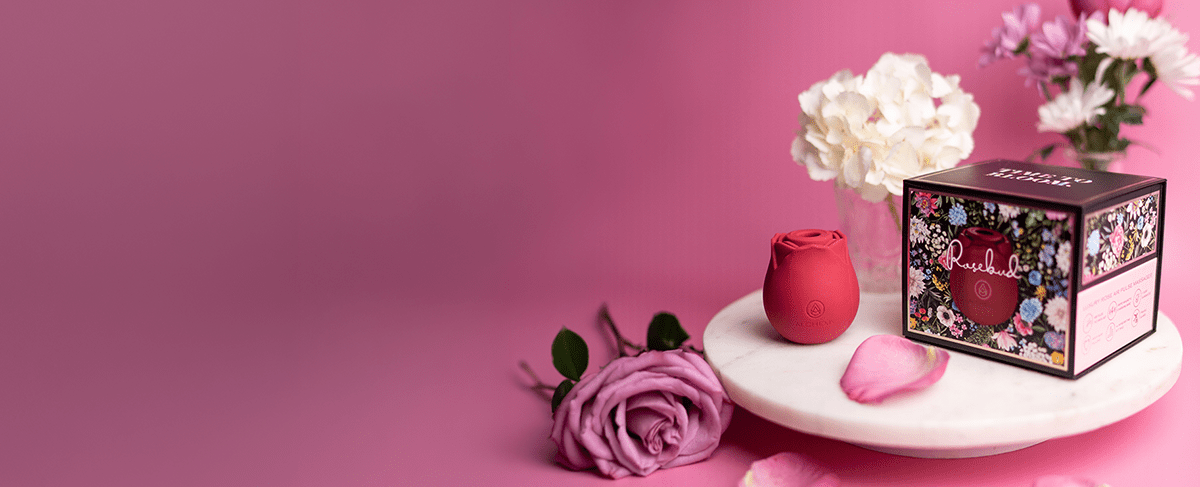 Alchemy Rosebud Luxury Air-Pulse Massager with its box on a round marble pedestal surrounded by roses and other flowers with a pink studio background