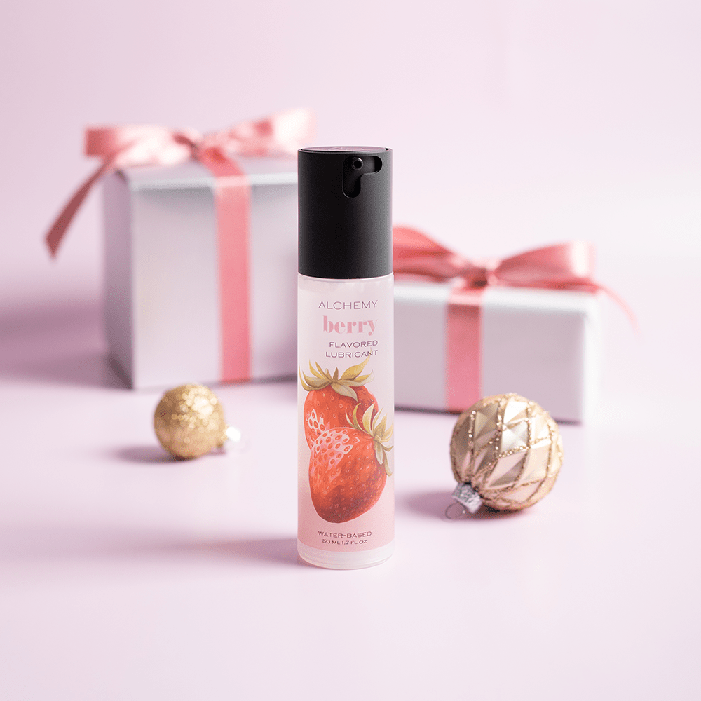 Alchemy Indulge: Strawberry Flavored Personal Lubricant bottle with holiday gift boxes, ornaments, and light pastel background