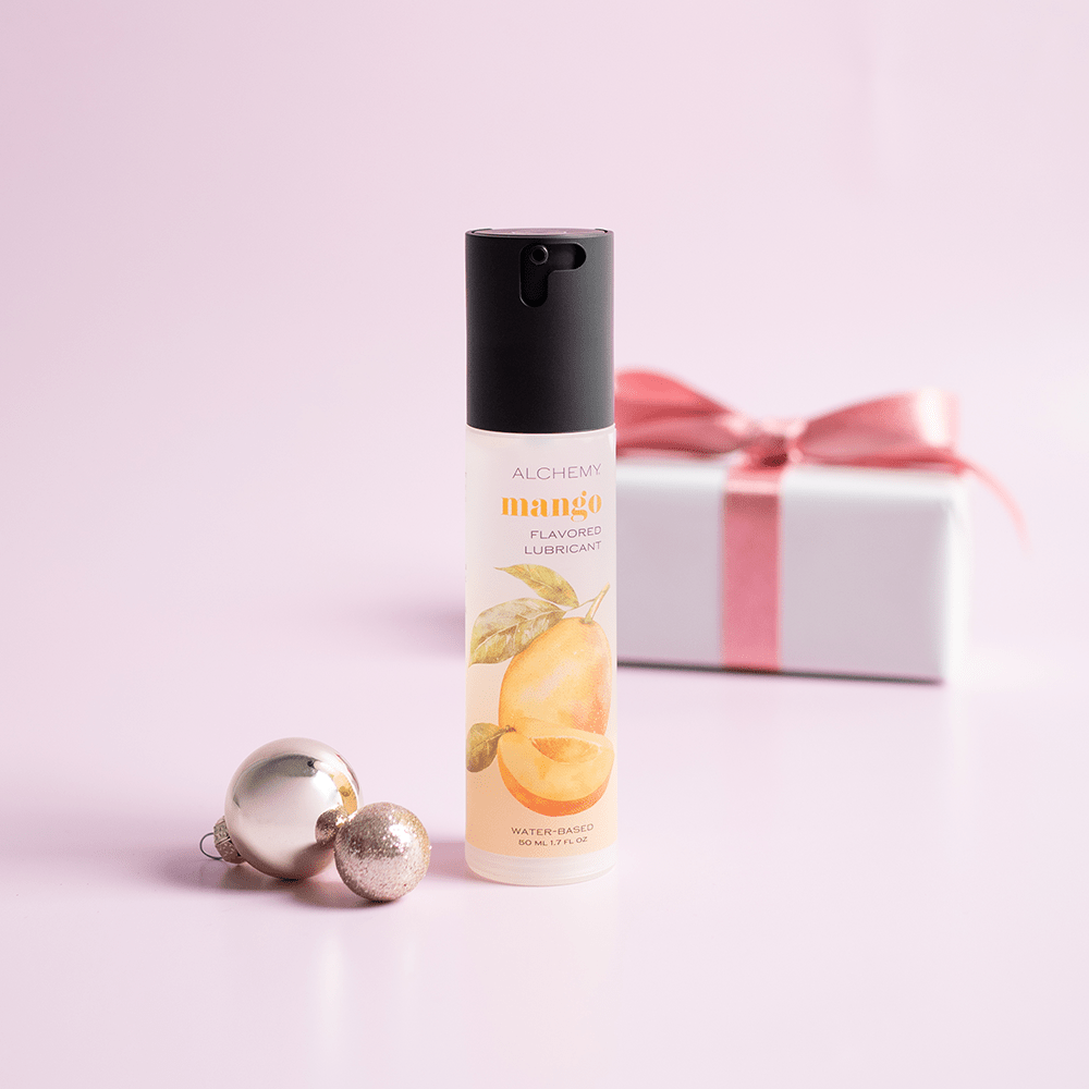 Alchemy Indulge: Mango Flavored Personal Lubricant bottle with holiday gift boxes, ornaments, and light pastel background