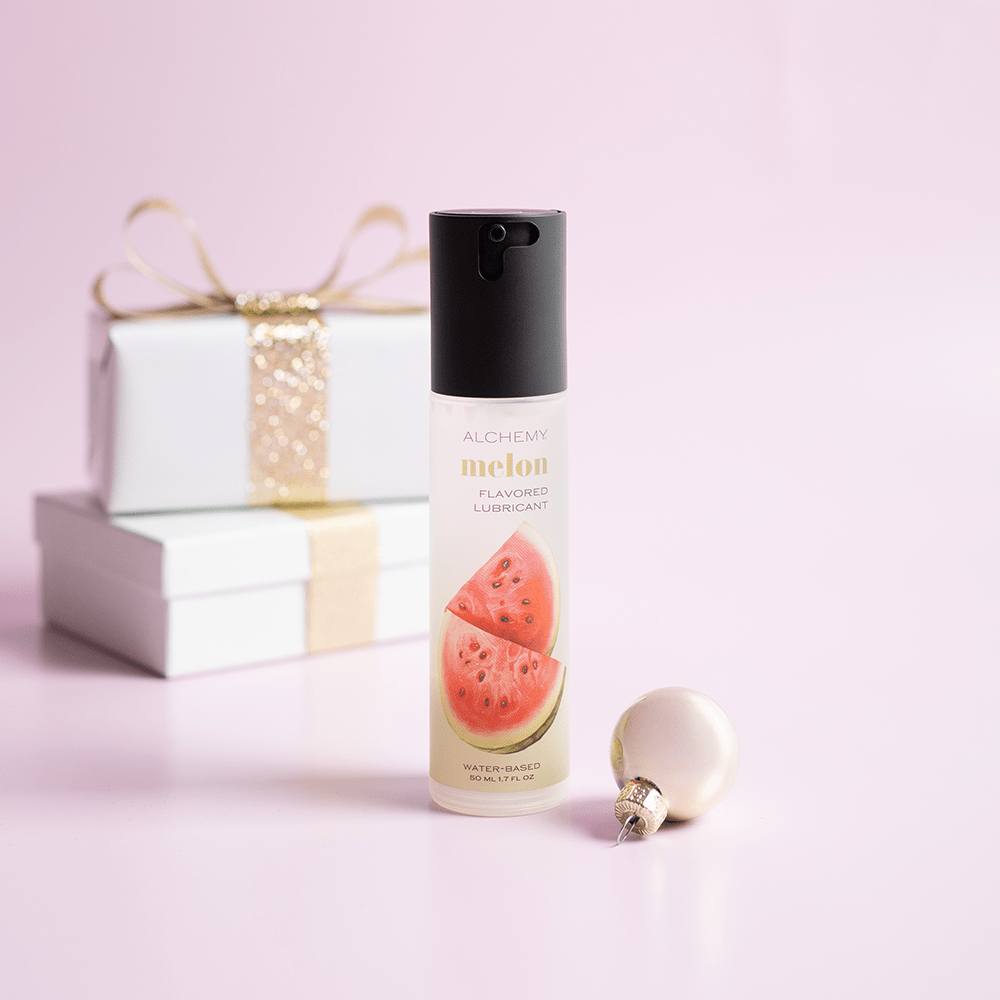 Alchemy Indulge: Watermelon Flavored Personal Lubricant bottle with holiday gift boxes, ornaments, and light pastel background