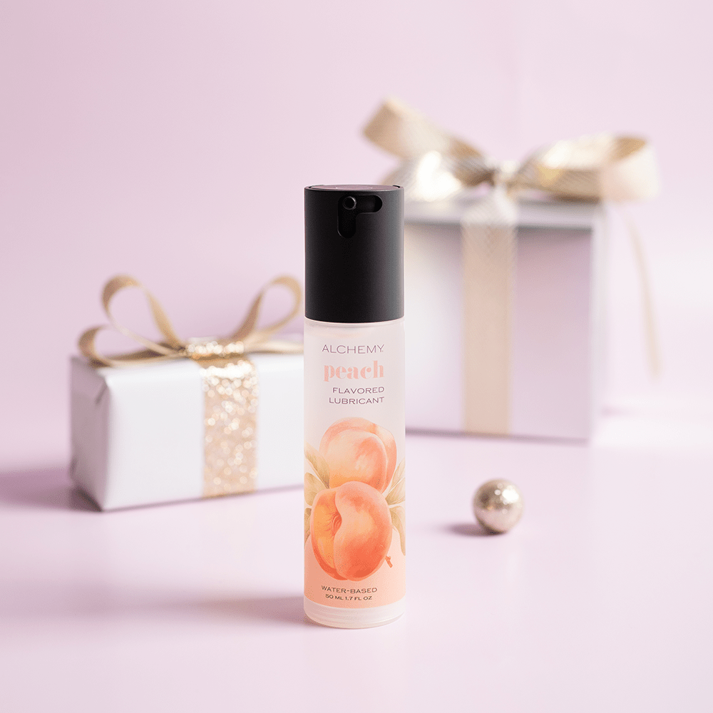 Alchemy Indulge: Peach Flavored Personal Lubricant bottle with holiday gift boxes, ornaments, and light pastel background