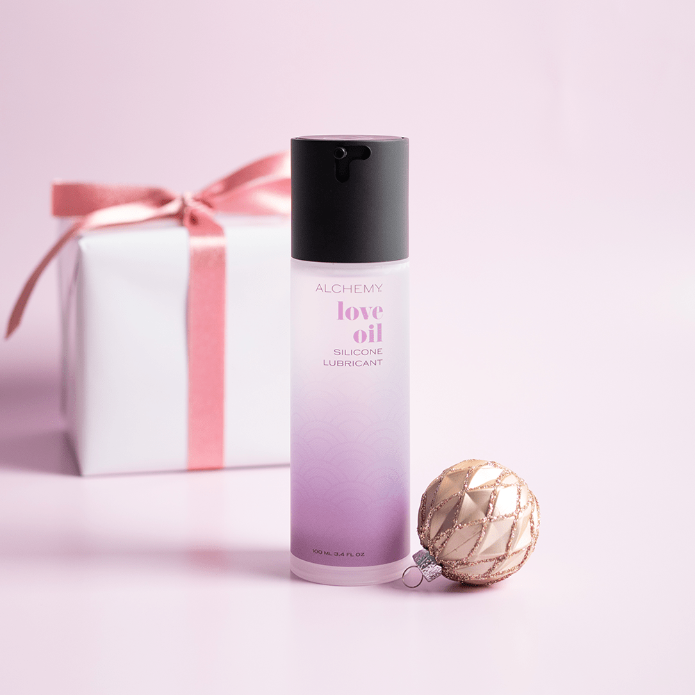 Alchemy Love Oil: Silicone-Based Personal Lubricant bottle with holiday gift boxes, ornaments, and light pastel background