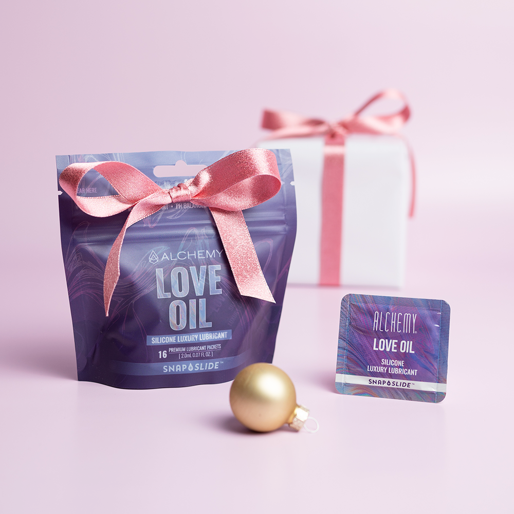 Alchemy Love Oil Silicone-Based Snap + Slide Single-Use Personal Lubricant Packets with holiday gift boxes, ornaments, and light pastel background