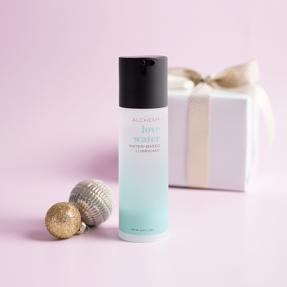 Alchemy Love Water: Water-Based Personal Lubricant bottle with holiday gift boxes, ornaments, and light pastel background