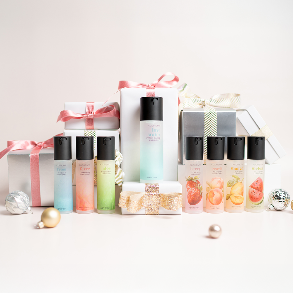 Alchemy Lush Trinity Bundle: Water-based personal lubricant bottle with holiday gift boxes, ornaments, and light pastel background
