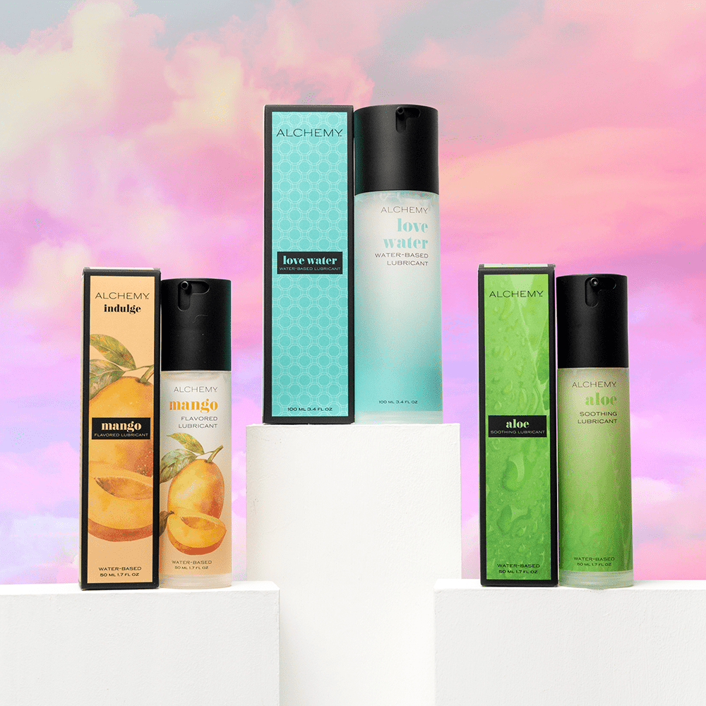 Alchemy Lush Trinity Bundle: Water-based personal lubricant bottles and boxes on white blocks and a colorful cloudy sky background