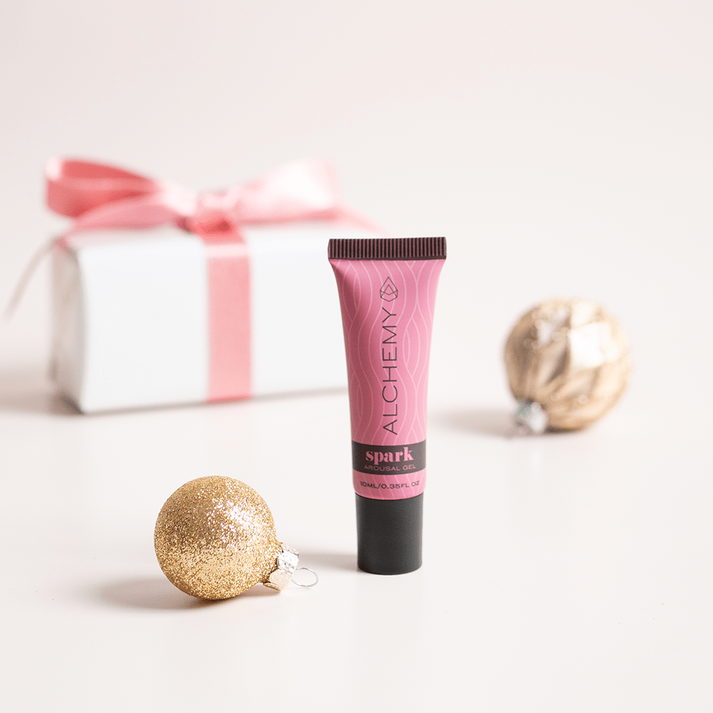 Alchemy Spark Female Arousal Gel 1oz tube with holiday gift boxes, ornaments, and light pastel background